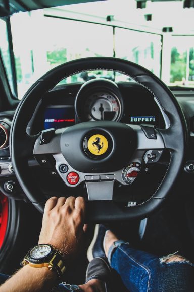Close-up of a Ferrari steering wheel with a hand and foot visible inside the car.
